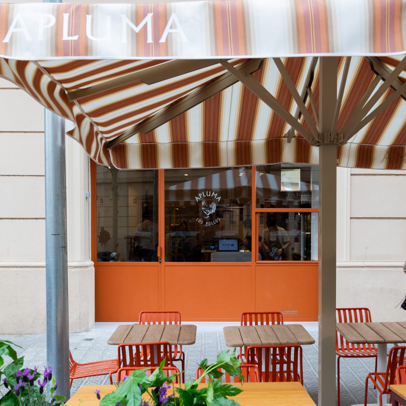 Fachada del restaurante APLUMA en Les Corts con carpintería y acceso en color naranja, toldo a rayas y terraza exterior.